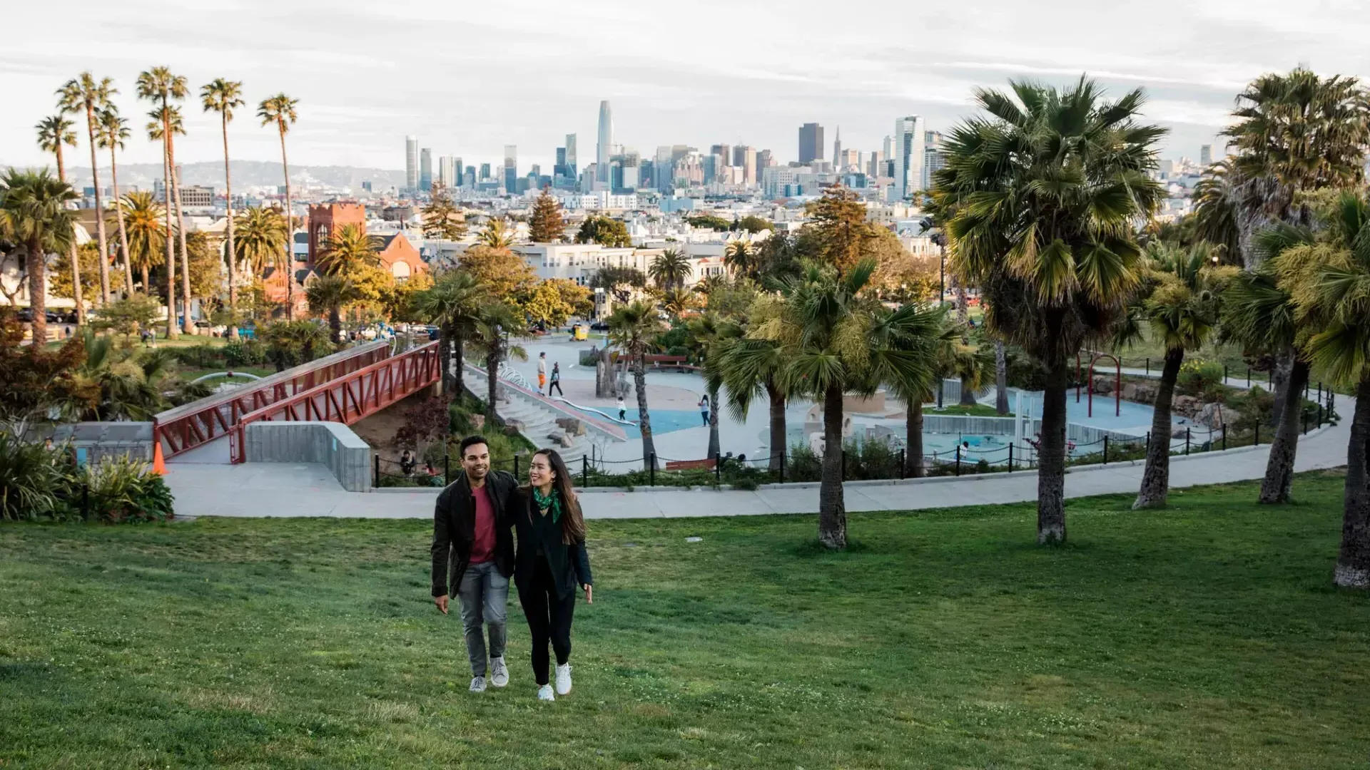 一对夫妇走向镜头，Dolores Park和威尼斯人官网平台app的天际线在他们身后.